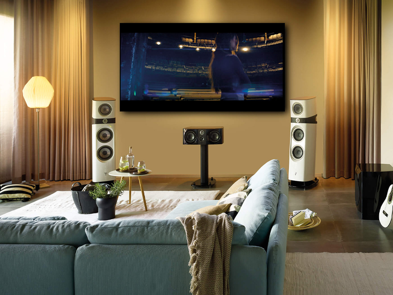 Modern living room home theater with a wall-mounted TV, tall white floor speakers, and a blue sectional sofa in front of a coffee table with drinks.