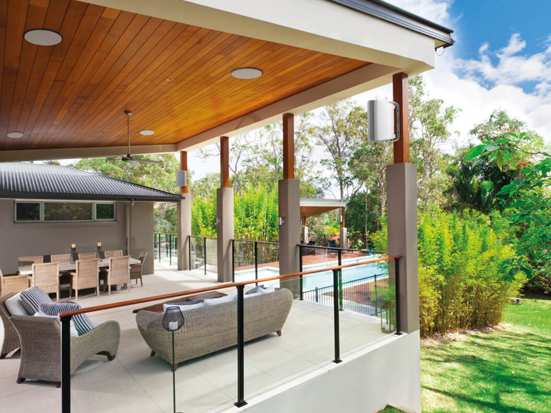 Covered outdoor patio with wicker seating, dining table, and glass railing overlooking a pool and lush garden area.