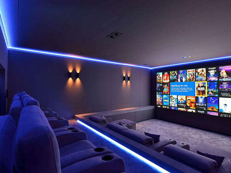 Private home theater with blue ambient lighting, plush gray recliner seats in rows, facing a large screen displaying movie posters.