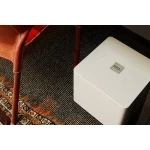 White rectangular, rounded-edge box resting on a dark patterned rug beside a brown leather chair leg and strap; a small metal plaque sits on its top surface.