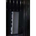 Tall white floor-standing speaker with multiple black drivers in a dim room, standing on small feet beside a textured curtain.