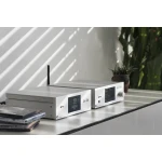 Two white hi‑fi audio units with digital displays on a desk, CDs to the left and a potted plant in the background.
